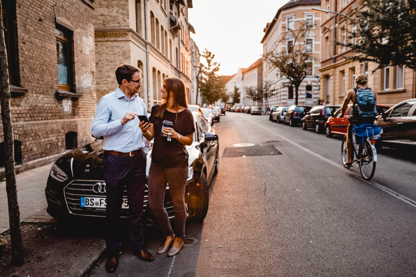 Homem e mulher conversando em frente a um carro estacionado na rua enquanto olham um celular. 