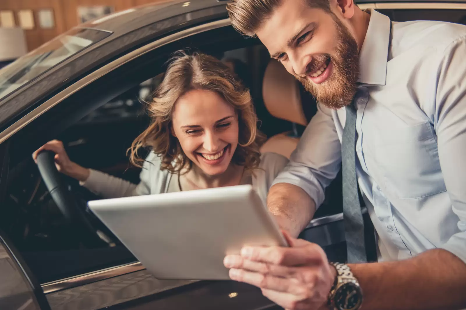 Homem mostrando tablet para mulher, que está dentro de um carro com as mãos no volante. Ambos sorriem. 