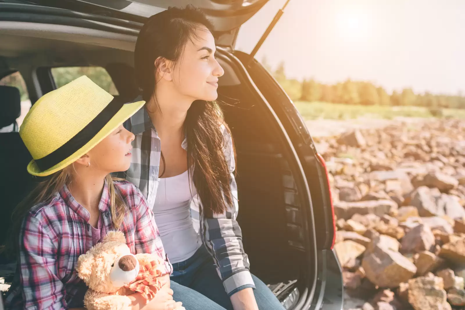 Seguro Garantia Estendida Volkswagen: mulher e criança sentadas no porta-malas de um carro olhando para o horizonte. A menina está usando um chapéu amarelo e segurando um ursinho de pelúcia. 