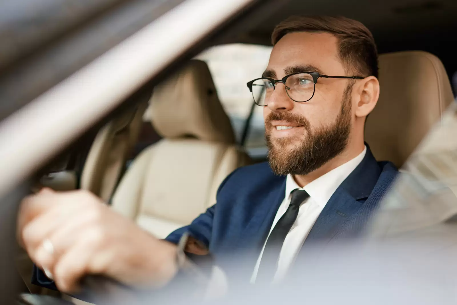 Foto de homem sorrindo e dirigindo um carro com bancos de couro na cor marrom. O homem possui barba e usa óculos de grau com armação preta. Veste blusa branca, terno azul e gravata preta. A imagem é captada da janela do motorista do carro.
